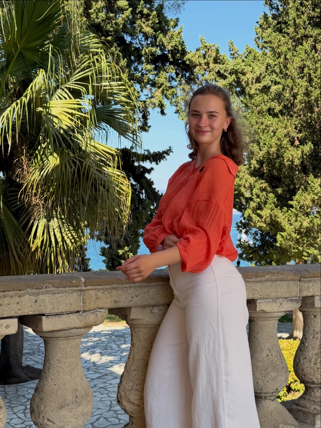 Sofiia Nosenko in a coral blouse beside a stone balustrade and sunlit trees